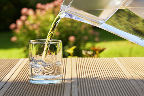 Pouring clear filtered water from a water filtration jug into a glass on the green summer garden background in a warm sunny summer day